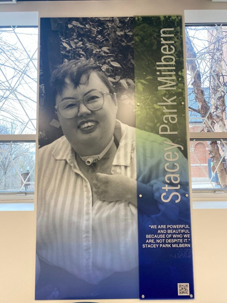 A black and white photo of Stacey Park Milbern, a mixed race Korean and white queer person, smiling while wearing a striped shirt and glasses. Milbern’s trach and wheelchair can be seen. To the right of the image, the text reads: “Stacey Park Milbern. We are powerful and beautiful because of who we are, not despite it. Stacey Park Milbern.” 