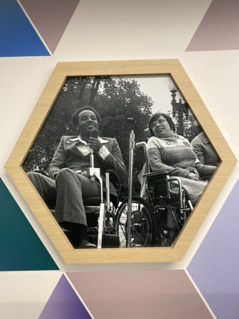 A black and white photo of Brad Lomax and Judy Heumann sitting in their wheelchairs next to one another while Brad holds a microphone. The photo is in the shape of a hexagon, with other geometric shapes around them. 