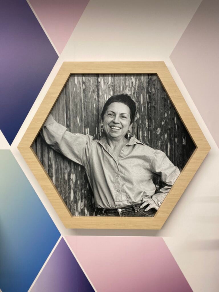 A black and white photo of Gloria Anzaldua, leaning against a wall and smiling. Braille and a QR code are below. The photo is in the shape of a hexagon, with other geometric shapes around them. 