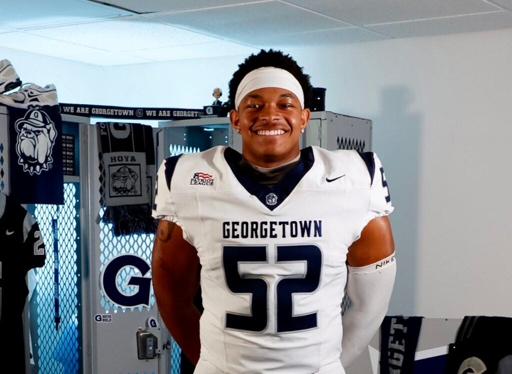 Smiling young man wearing a white Georgetown football jersey with the number 52, standing in a team locker room.