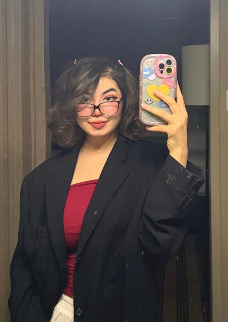Selfie of a young woman with short wavy hair and glasses wearing a black blazer over a red top, holding up a decorated phone case.