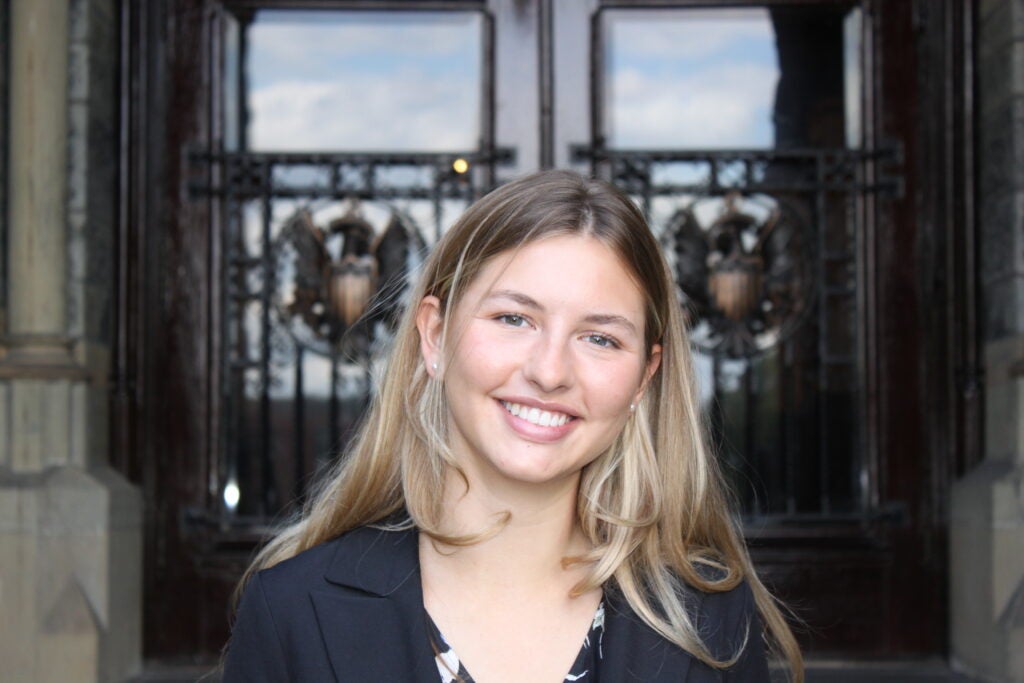 Portrait of a smiling young woman with blonde hair, wearing a black blazer in front of decorative glass and metal doors.