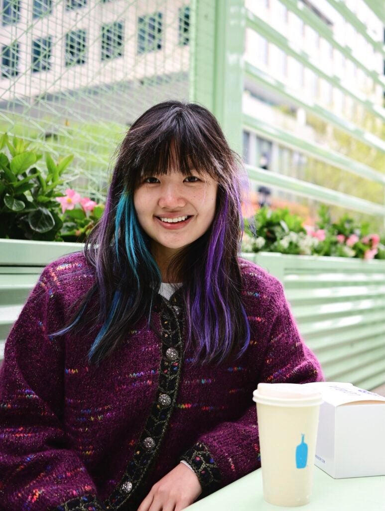 Young woman with purple and blue highlights in her hair, wearing a purple knit cardigan, sitting outdoors at a table with a coffee cup.