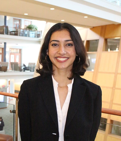 Professional headshot of a smiling woman with shoulder-length dark hair, wearing a black blazer and white top inside an office atrium.