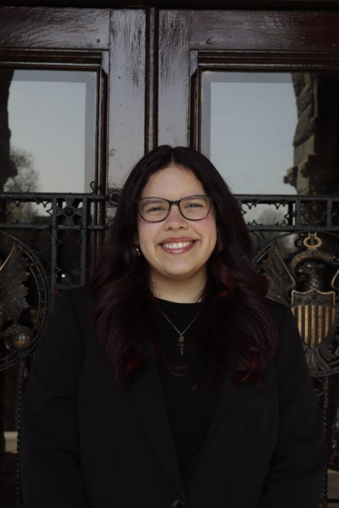 Portrait of a smiling woman with dark red hair and glasses, wearing a black blazer in front of ornate glass doors.