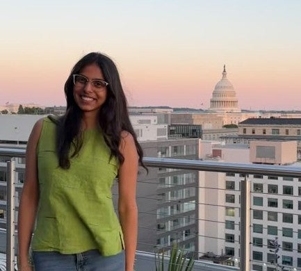 Smiling woman with long dark hair and glasses in a green sleeveless top, standing on a rooftop with the U.S. Capitol dome in the background.