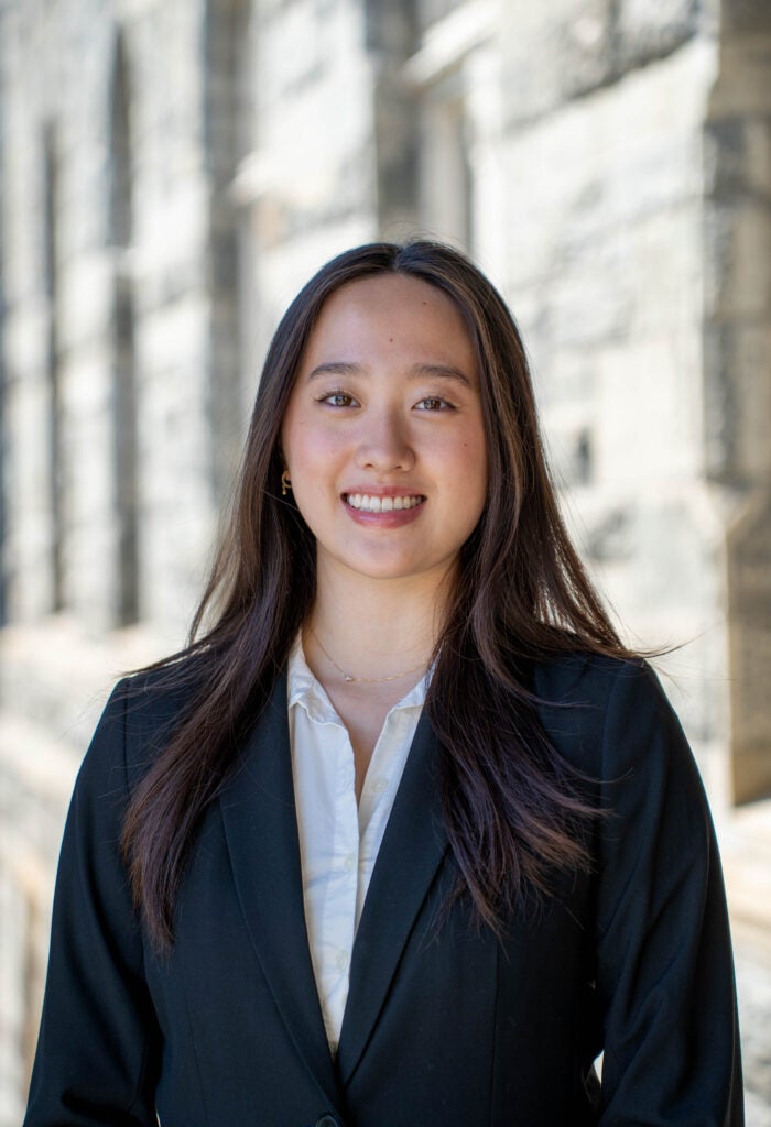 Professional headshot of a smiling young woman with long dark hair, wearing a black blazer over a white shirt against a stone background.