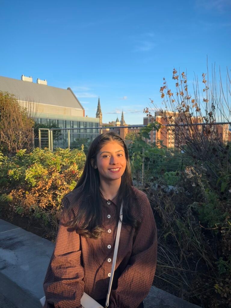 Young woman with dark hair in a brown patterned shirt smiling outdoors on a rooftop garden with city buildings behind her.