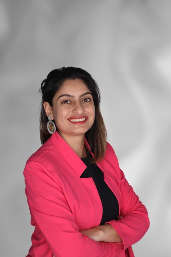 Professional portrait of a smiling woman wearing a bright pink blazer over a black top, standing with her arms crossed against a light gray background.